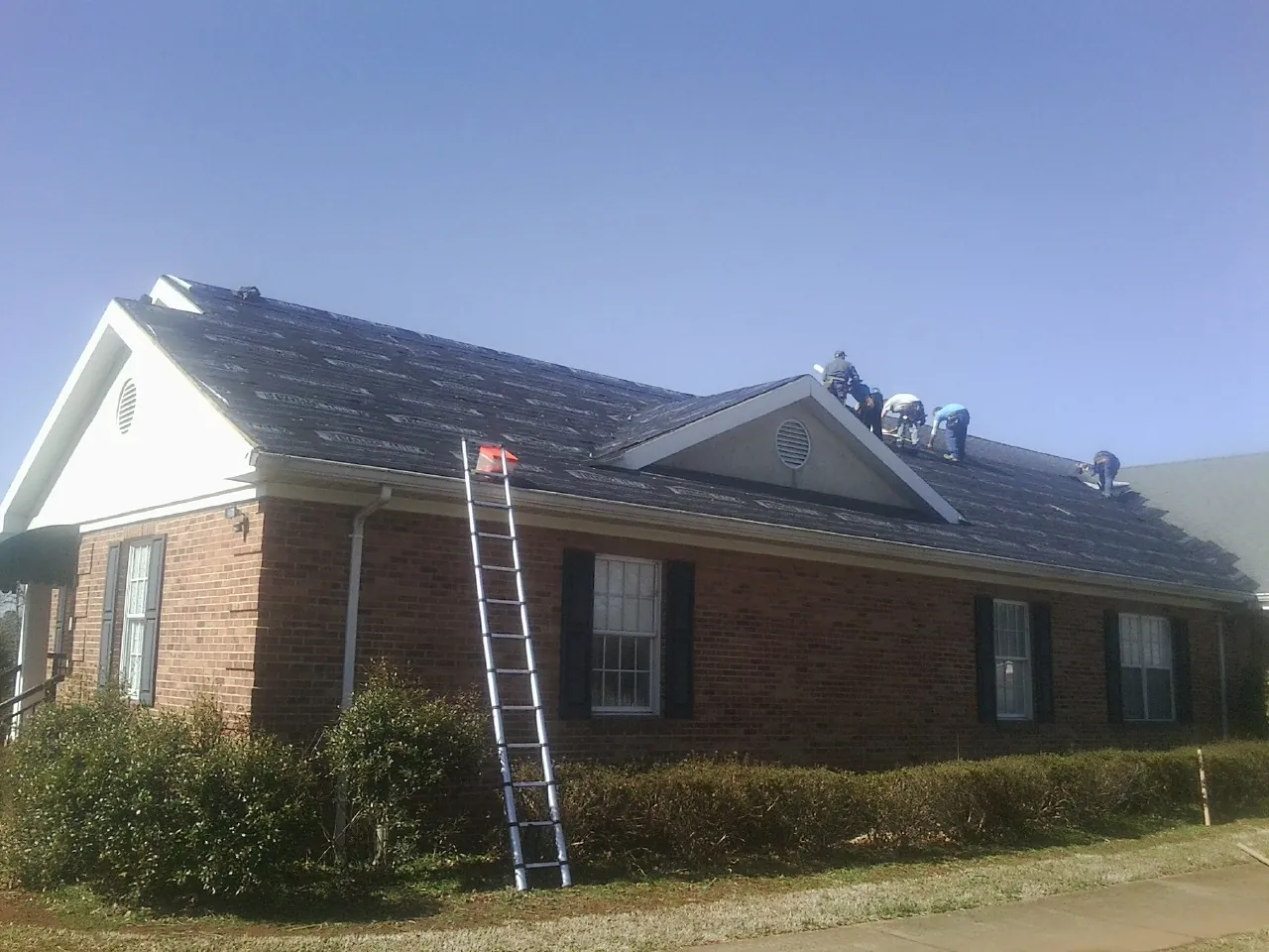 Crew tearing shingles off a brick ranch roof with a ladder set — Emergency Roof Repair work by our crew in Chokio