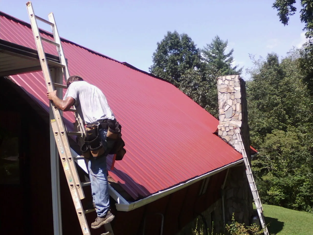 Roofer on a ladder beside a red metal roof and stone chimney — Soffit & Fascia Repair by Roofing Brothers in Chokio